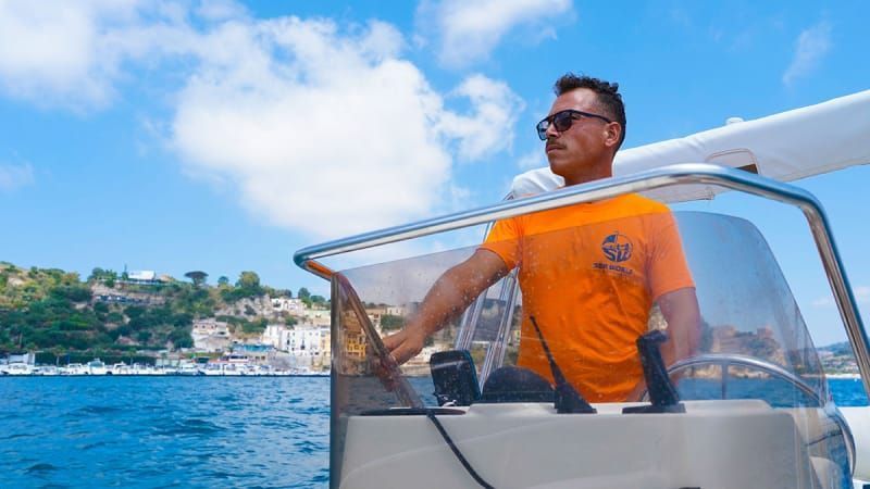 Man steering a boat on a sunny day, wearing sunglasses and an orange shirt, with a coastal view in the background.