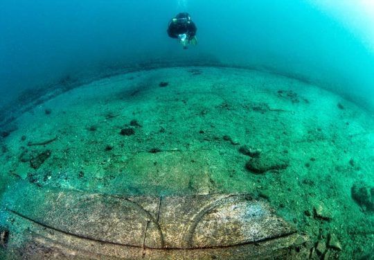 Diver swims above large, rounded underwater structure with curved markings. Turquoise water.