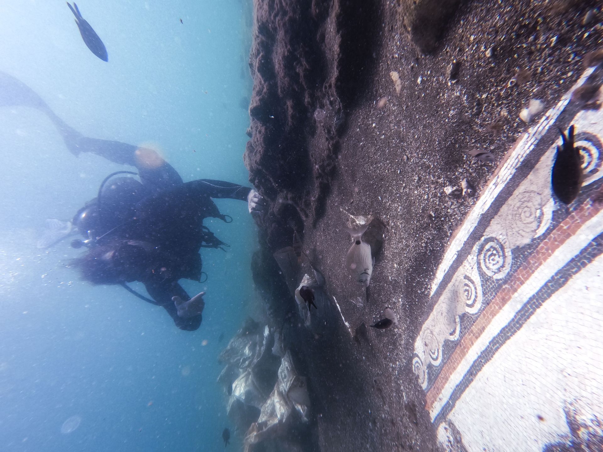 Diver examines underwater ancient wall with painted decorative patterns. Marine life swims nearby.