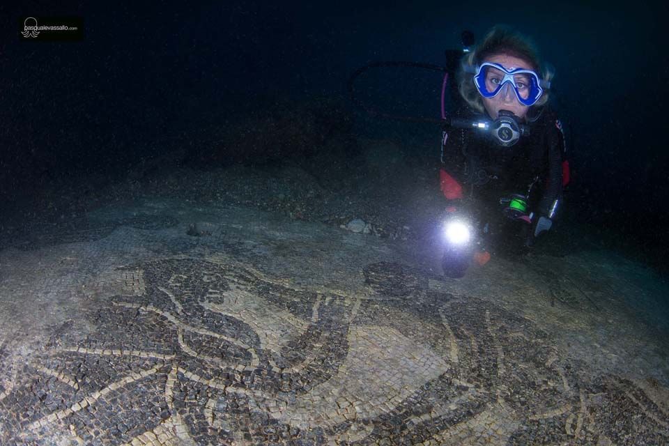 Diver with flashlight examines submerged petroglyphs on a dark seafloor.