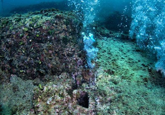 Underwater view of rocky seabed with air bubbles, a section of what appears to be a structure, and debris.