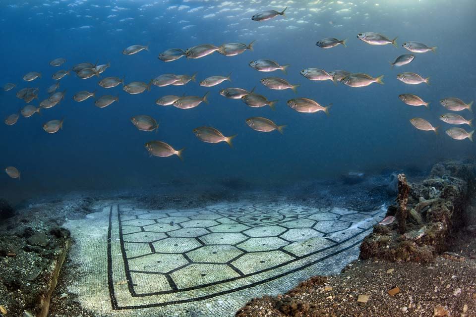 Underwater view of a mosaic floor with a school of fish swimming above it in blue water.