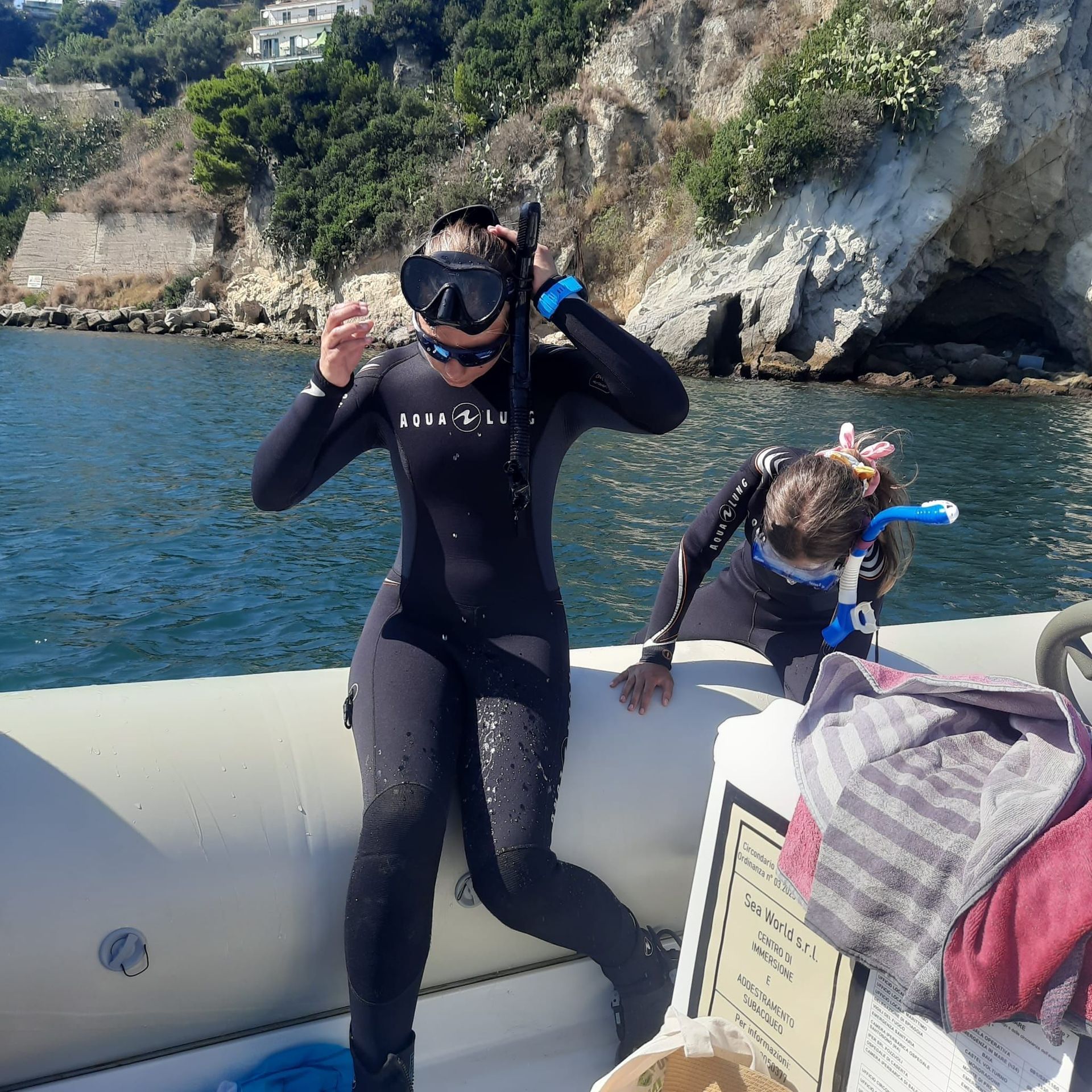 Two people in wetsuits prepare to snorkel on a boat, against a coastal backdrop.