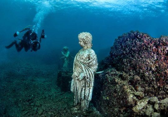 Underwater view of ancient statues; diver explores. Statues are white, coral and deep blue water.