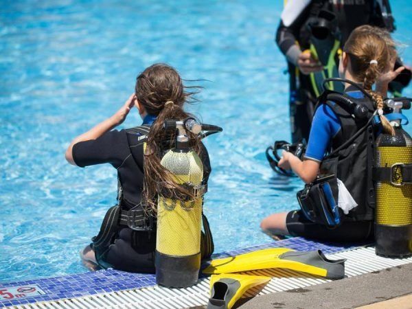 People in scuba gear preparing to enter pool. One adjusting mask, others sitting. Yellow tanks and fins visible.