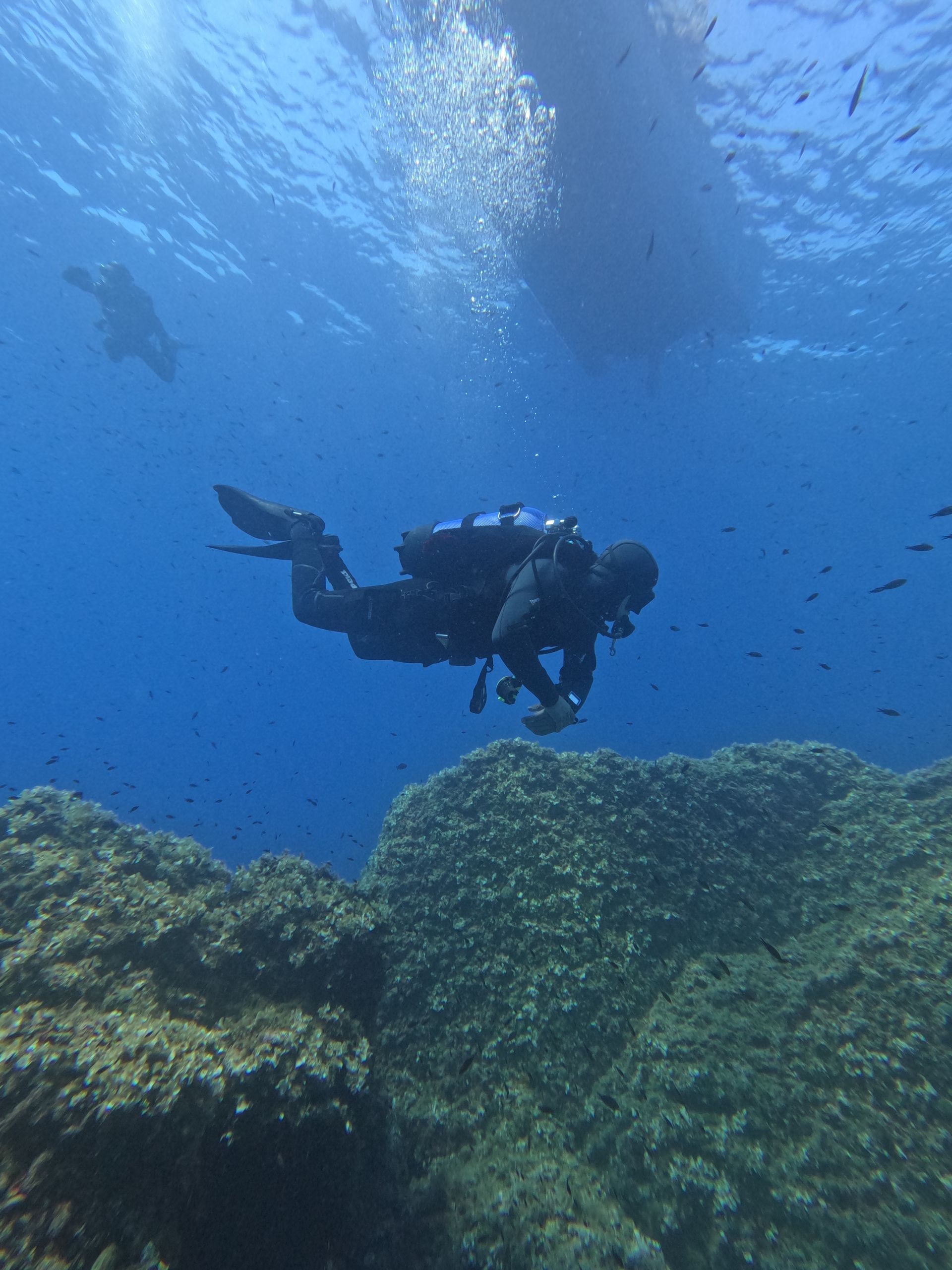 Diver explores a rocky underwater environment, boat above. Blue water, dark wetsuit, bubbles, and sunlight.