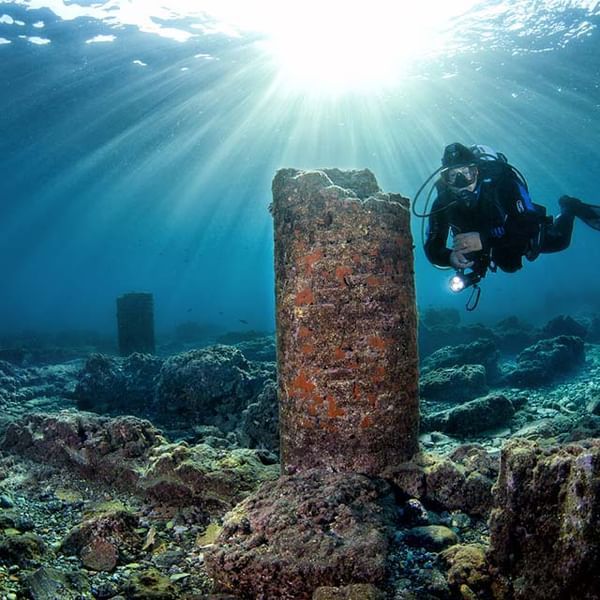 Scuba diver exploring underwater ruins, sun shining through the water; columns and rocks visible.