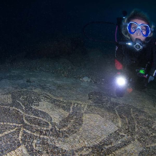 Diver underwater, illuminating ancient mosaic on sea floor. Dark water, person wearing scuba gear and goggles.