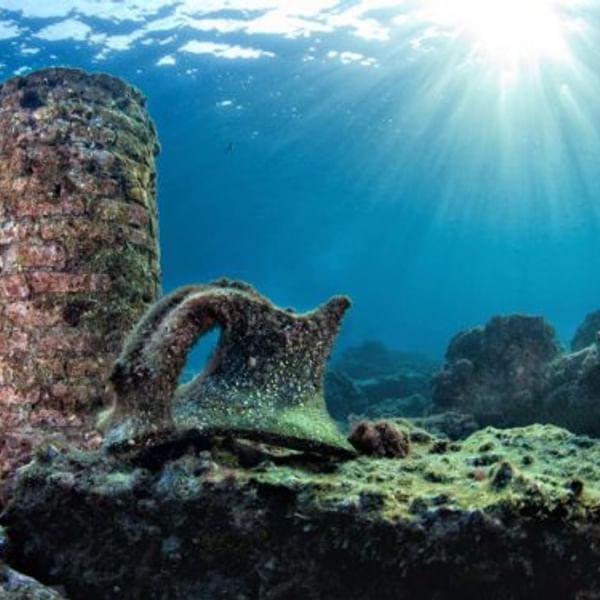 Underwater ruins, possibly ancient, with an aged jar on the ocean floor and sunlight filtering through the water.