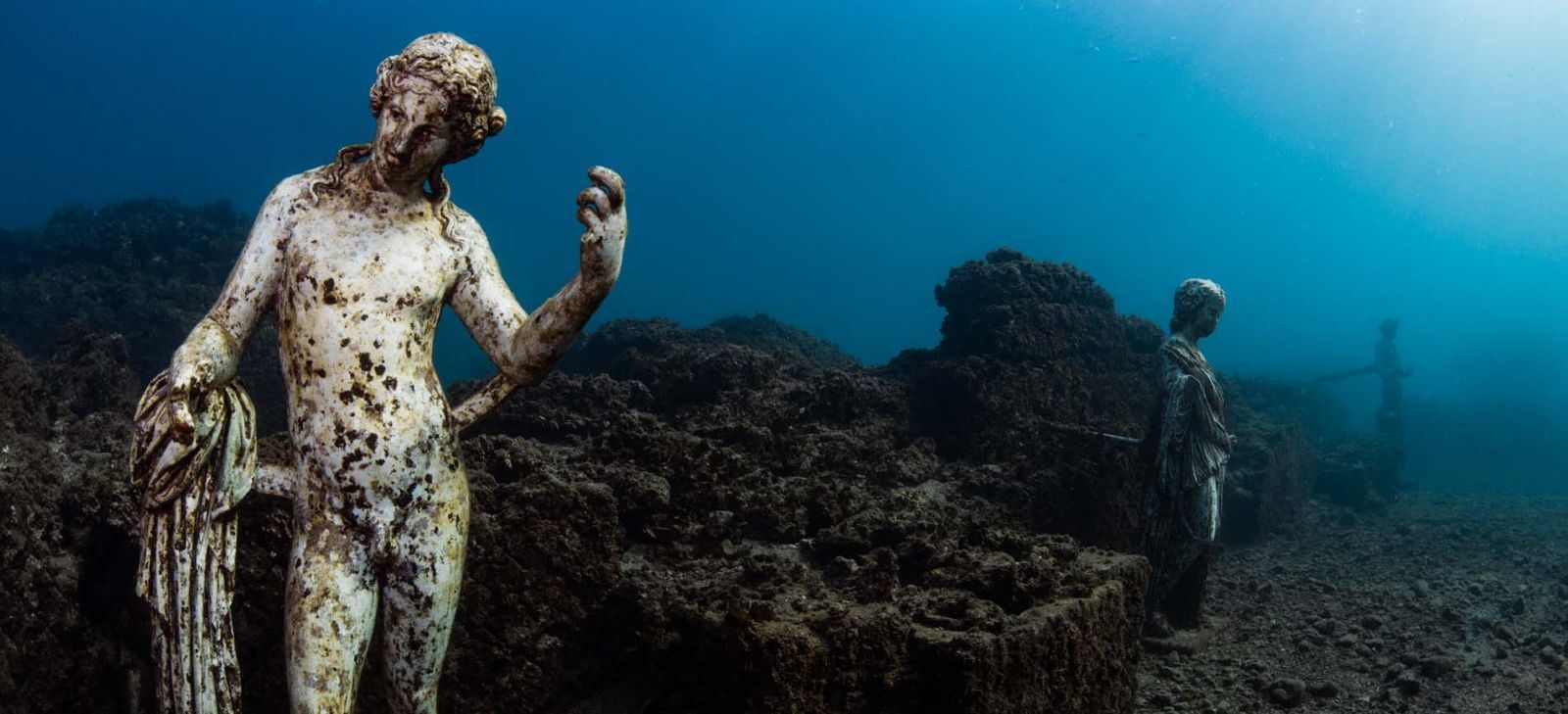 Statues submerged underwater, covered in algae. Clear blue water and dark seabed.
