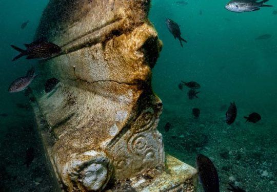 Underwater view of an ancient statue covered in algae, surrounded by schools of fish.