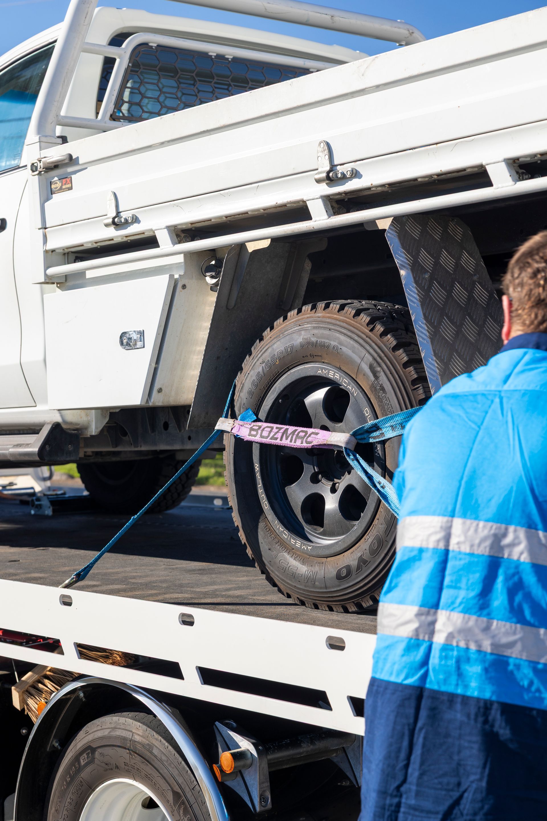 Person securing a white truck's tire to a tow truck with a blue strap, outdoors, sunny.— Perlyn Towing in Gladstone, QLD