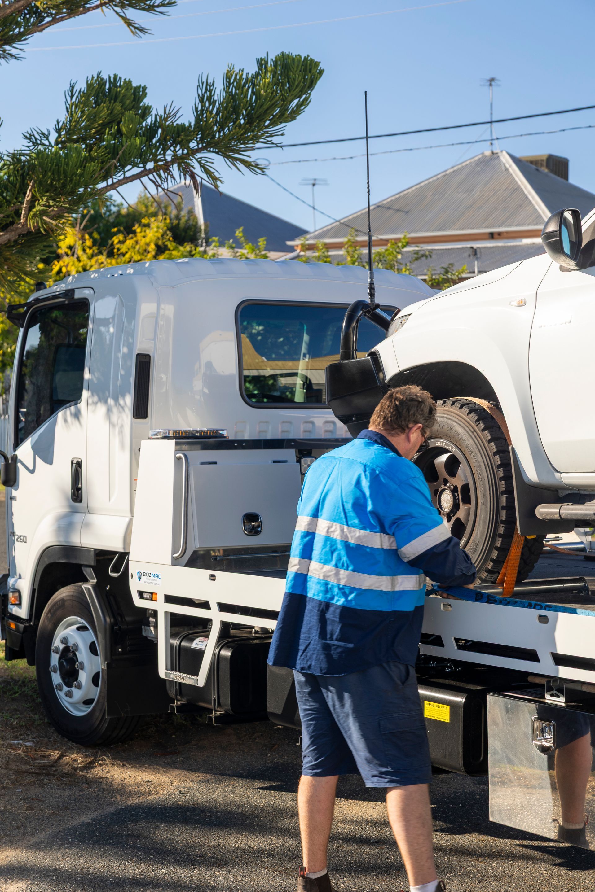A tow truck operator in blue and white work clothes secures a white vehicle on a tow truck in front of a house.— Perlyn Towing in Rockhampton City, QLD