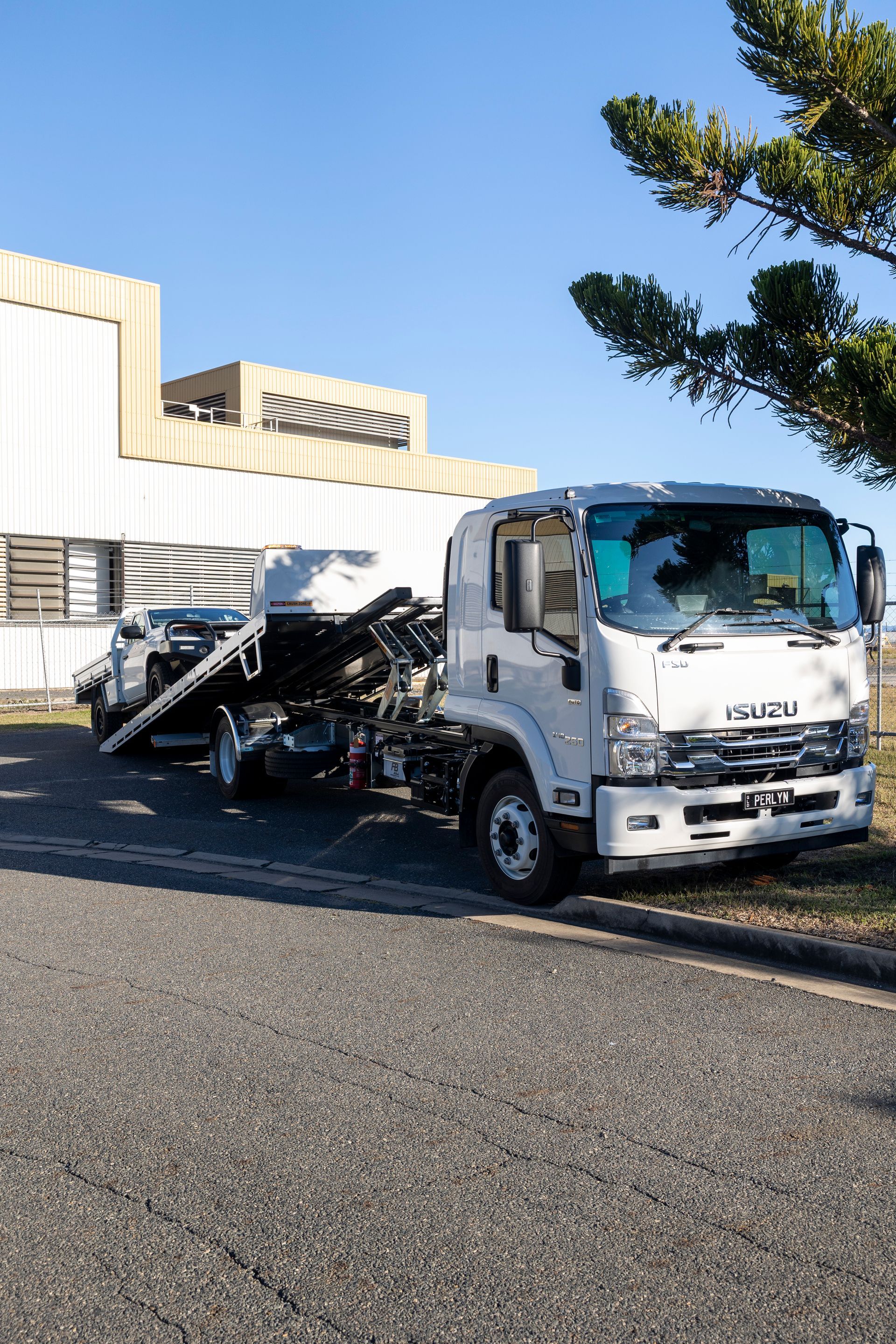 White Isuzu tow truck with a damaged car on its flatbed; parked on a paved area beside a building.— Perlyn Towing  in Emerald, QLD