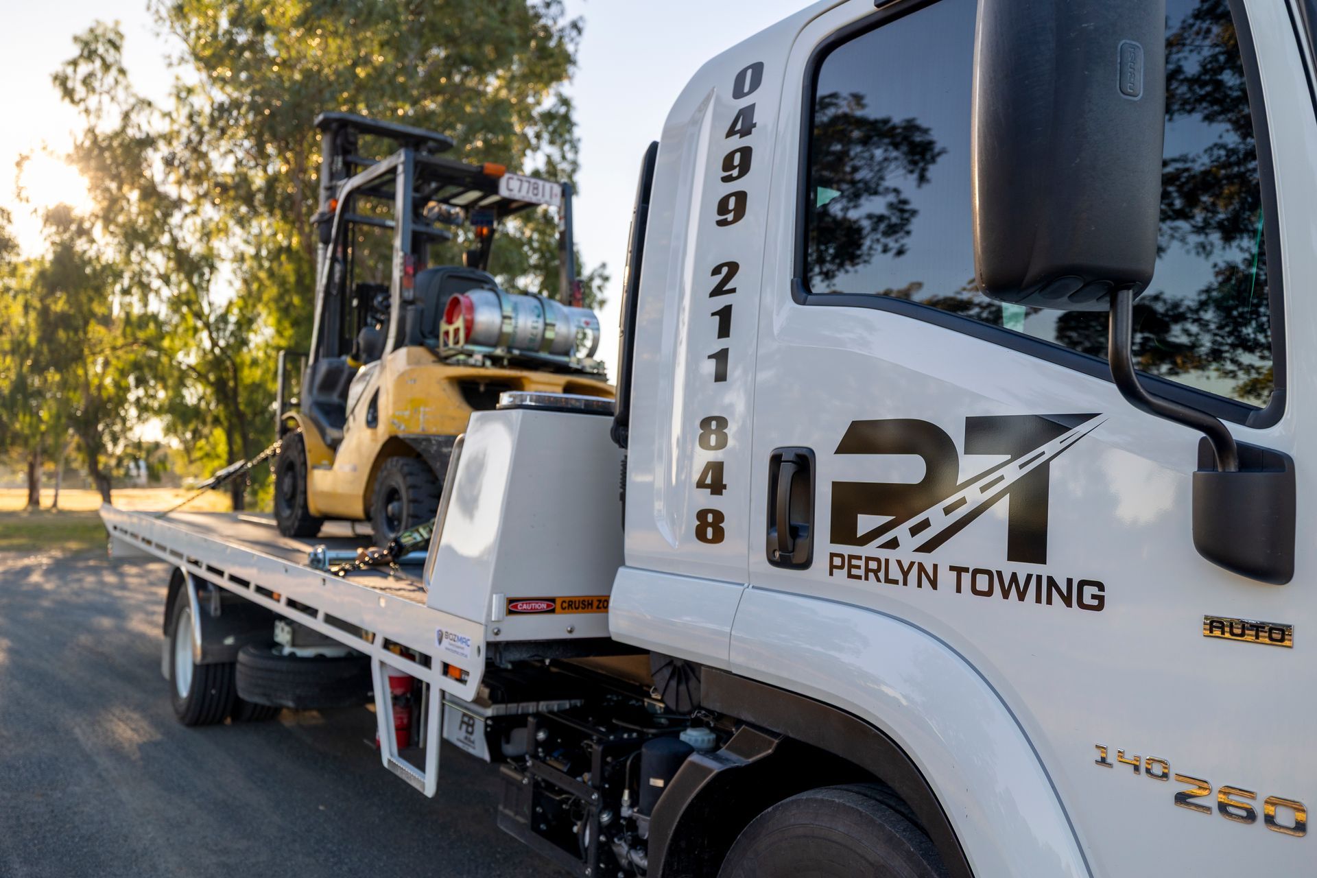 A white tow truck transporting a yellow forklift on its flatbed, with the sun setting in the background.— Perlyn Towing in Rockhampton City, QLD