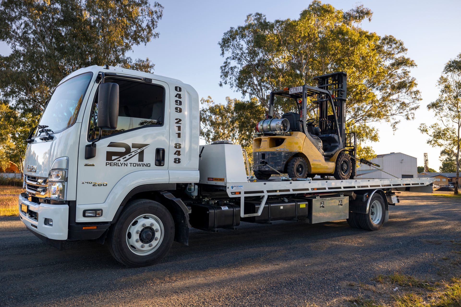White truck carrying a yellow forklift on a flatbed, outdoors.— Perlyn Towing in Gladstone, QLD