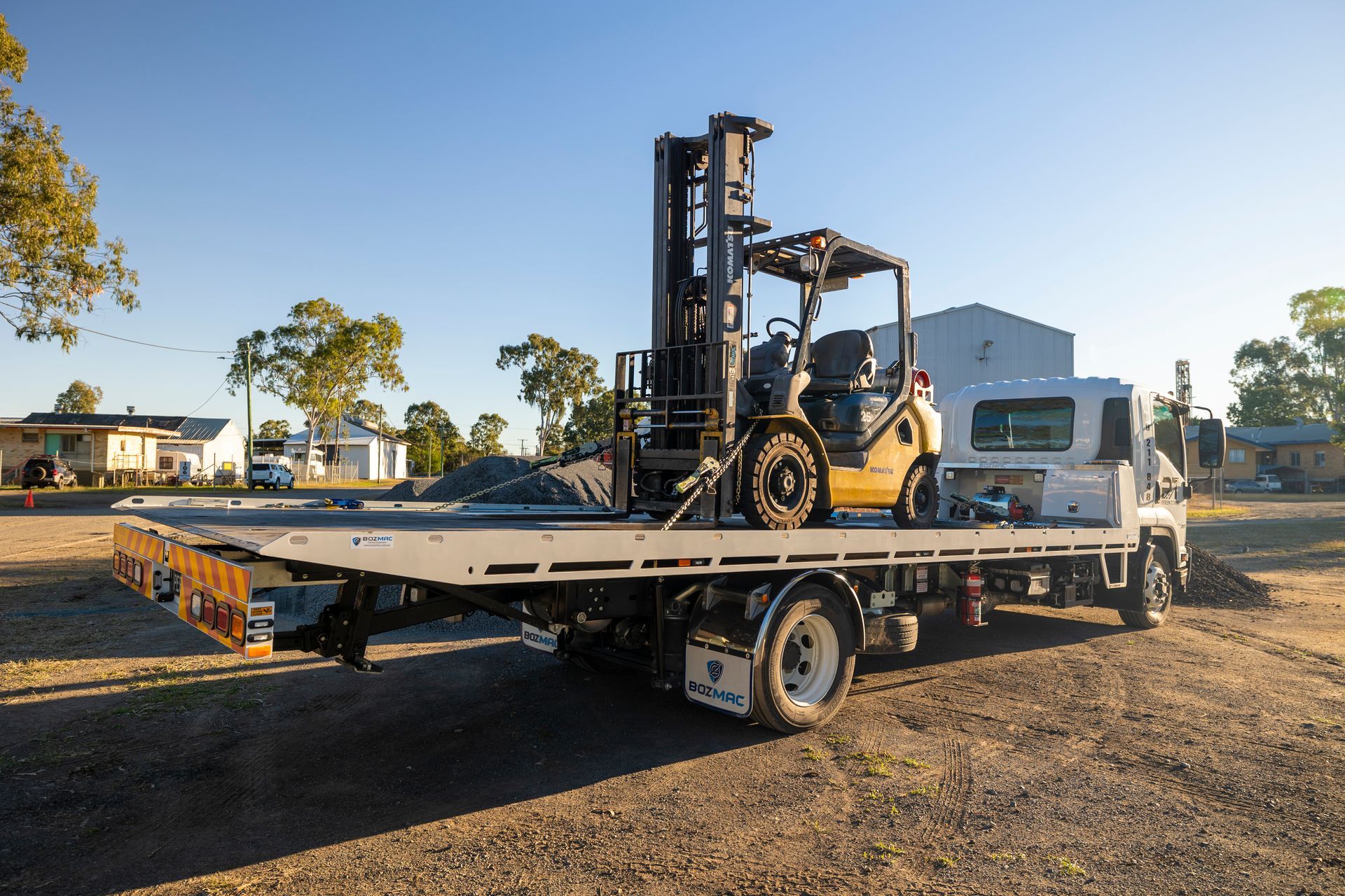 A flatbed truck with a yellow forklift on its bed, parked on gravel.— Perlyn Towing in Gladstone, QLD