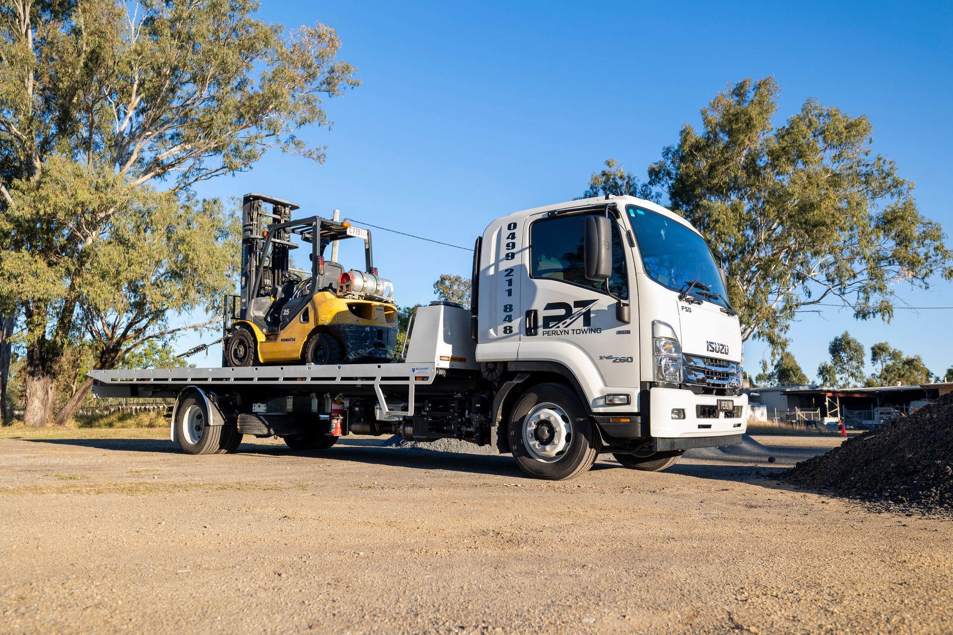 White Isuzu flatbed truck carrying a yellow forklift on a gravel lot under a blue sky.— Perlyn Towing  in Rockhampton City, QLD