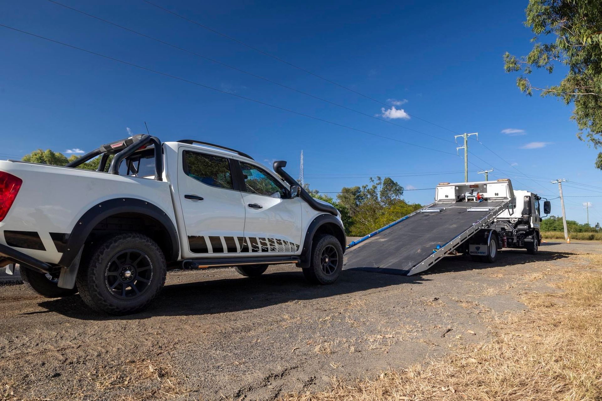 White pickup truck towing a flatbed tow truck on a dirt road under a blue sky.