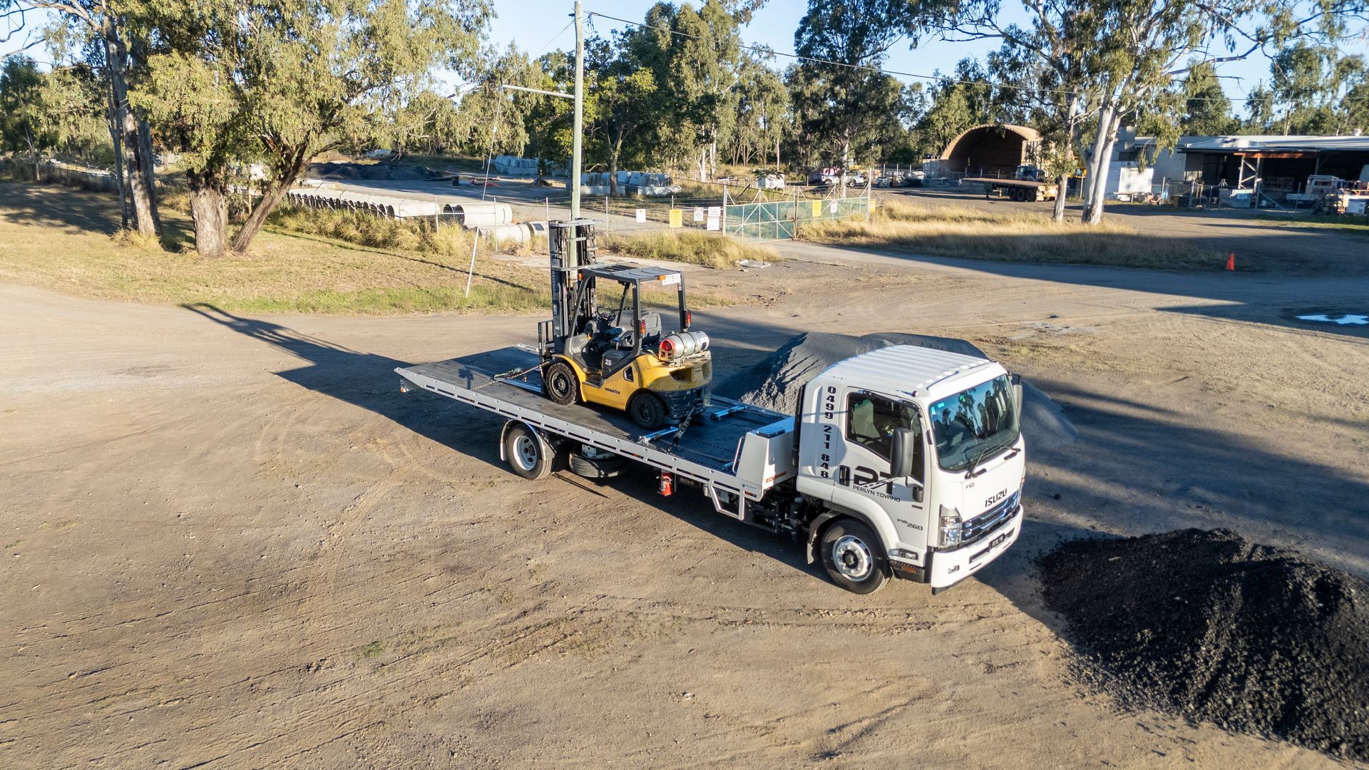 A flatbed truck carrying a yellow forklift on a gravel lot.— Perlyn Towing  in Rockhampton City, QLD