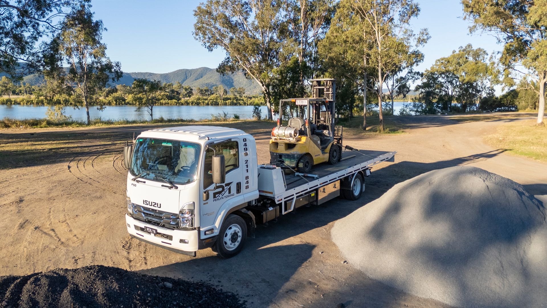 Flatbed truck carrying a forklift, parked on gravel, near a river.— Perlyn Towing  in Rockhampton City, QLD
