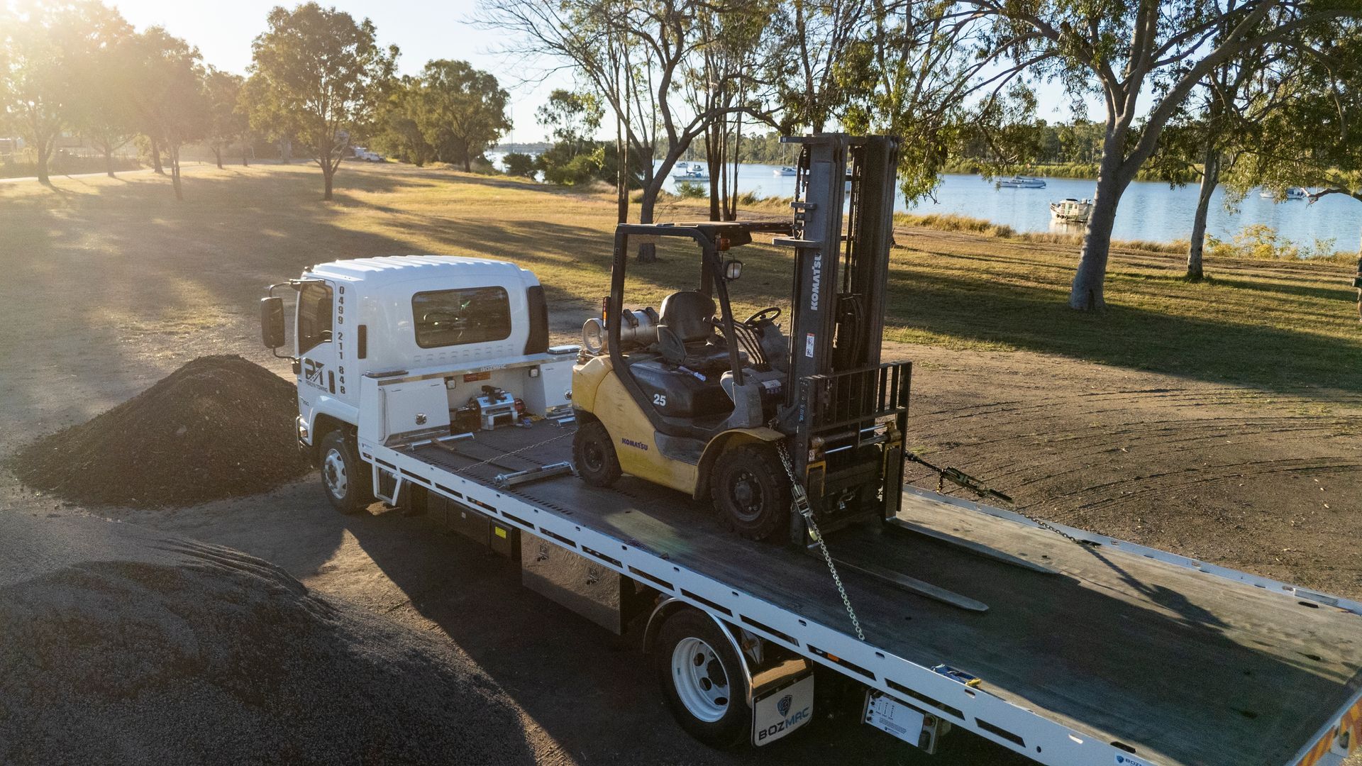 Yellow forklift on a flatbed truck near a body of water, with trees in the background.— Perlyn Towing  in Rockhampton City, QLD