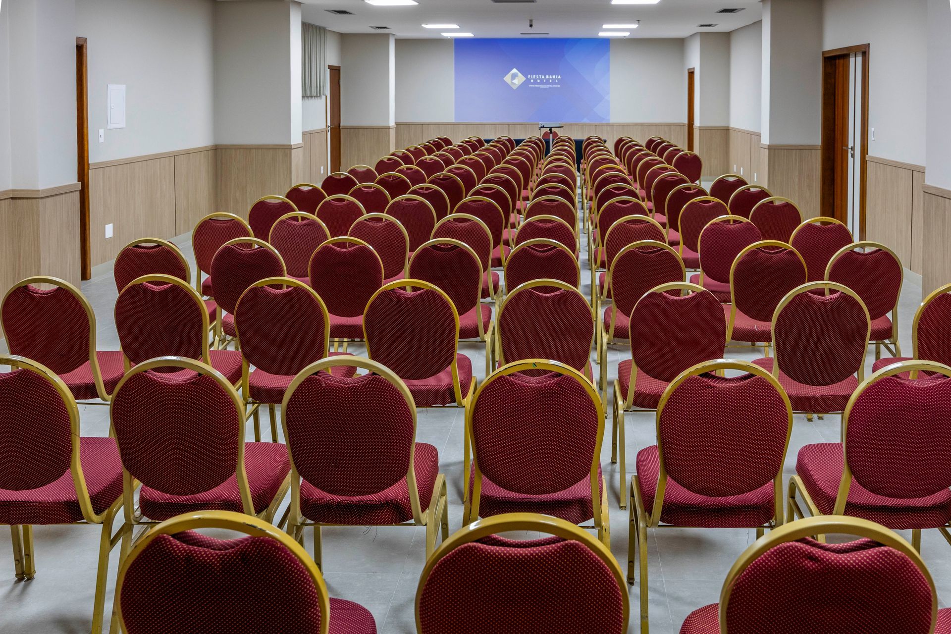 Uma sala de conferências com fileiras de cadeiras vermelhas e uma tela de projetor.