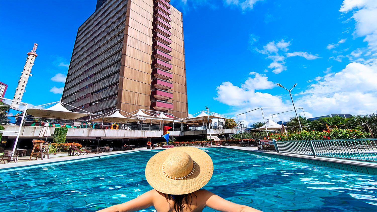 Uma mulher com um chapéu de palha está nadando em uma piscina em frente a um prédio alto.