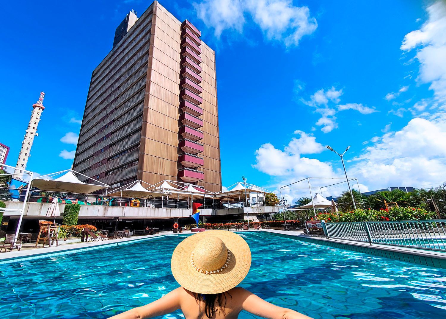 Uma mulher com um chapéu de palha está nadando em uma piscina em frente a um prédio alto.