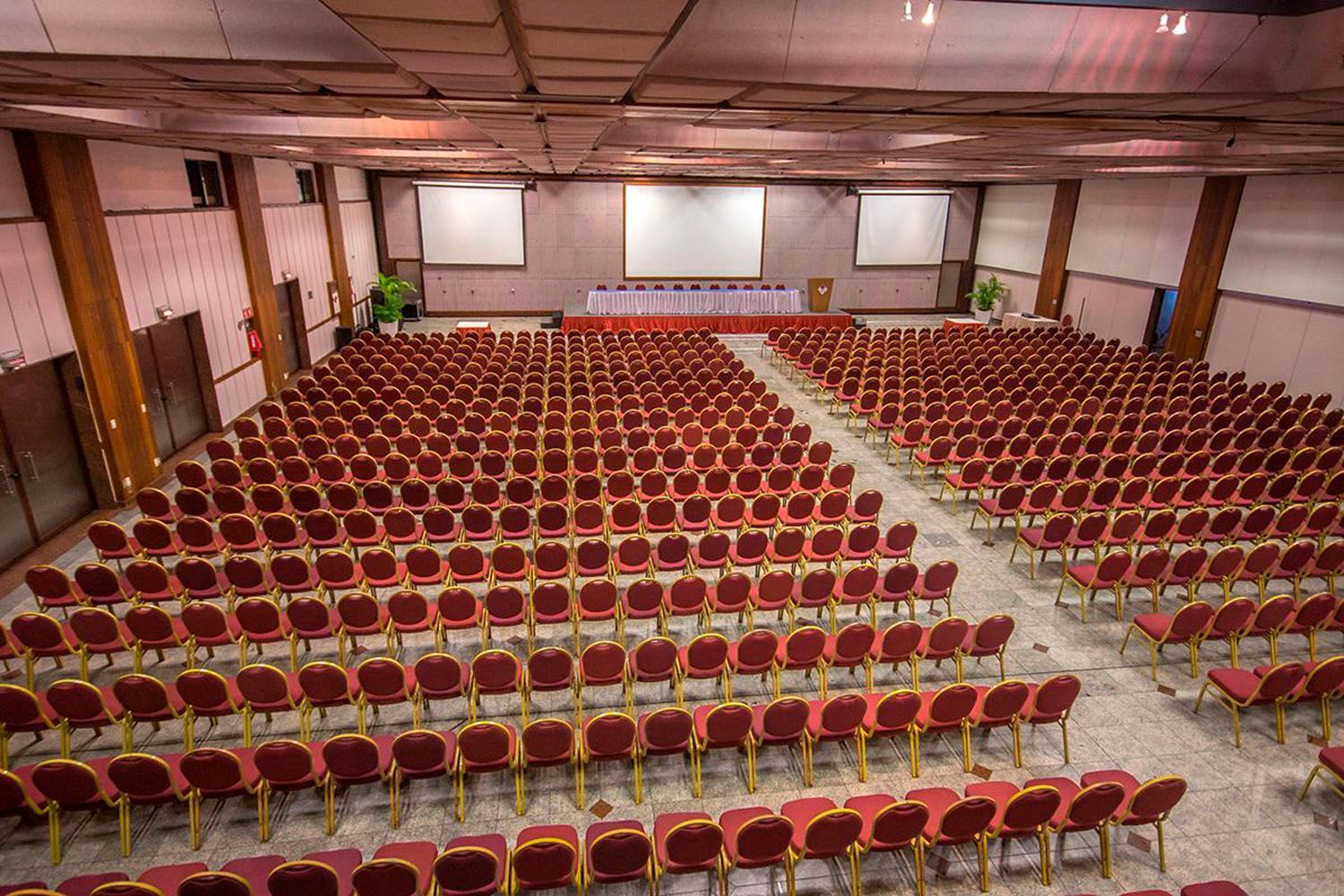 Uma grande sala de conferências com fileiras de cadeiras vermelhas e uma tela grande.