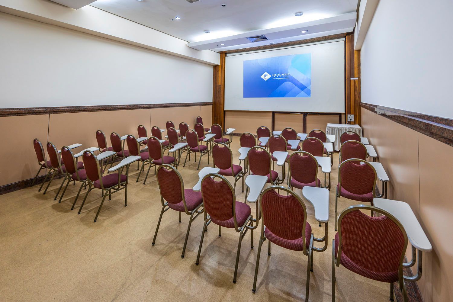 Uma sala de conferências com fileiras de cadeiras e mesas e uma tela de projetor.