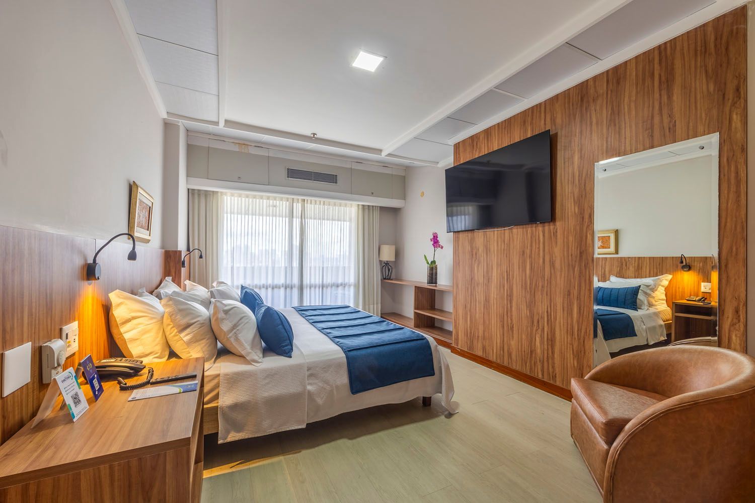 Um quarto de hotel com uma cama grande, cadeira, televisão e espelho.