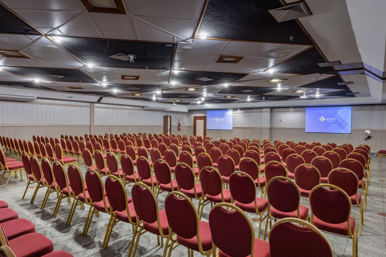 Uma grande sala de conferências com fileiras de cadeiras vermelhas e uma tela de projetor.