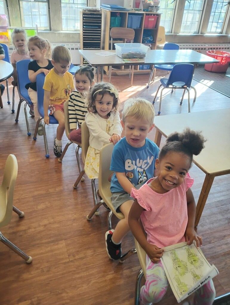 Children sitting in a row, looking towards the camera. Classroom setting.
