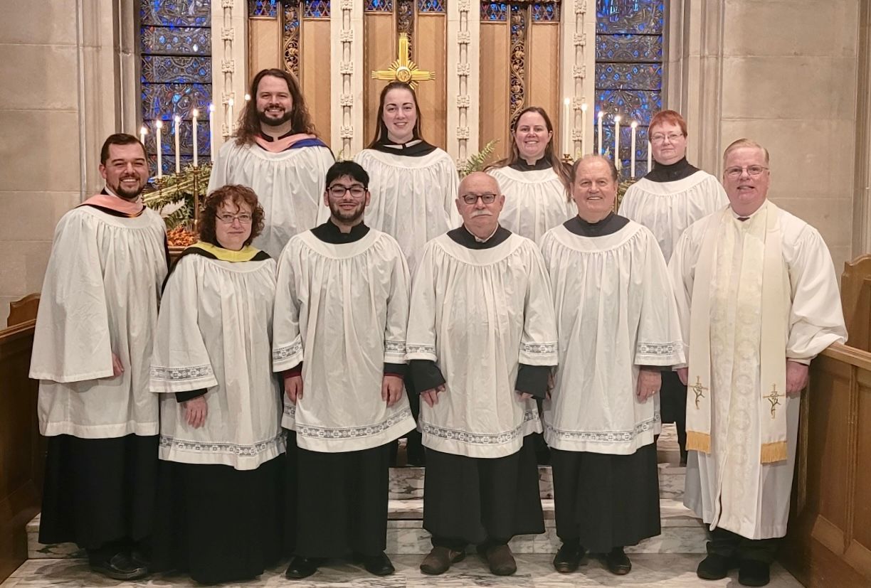 Choir group wearing white robes and black cassocks, standing in front of an altar.