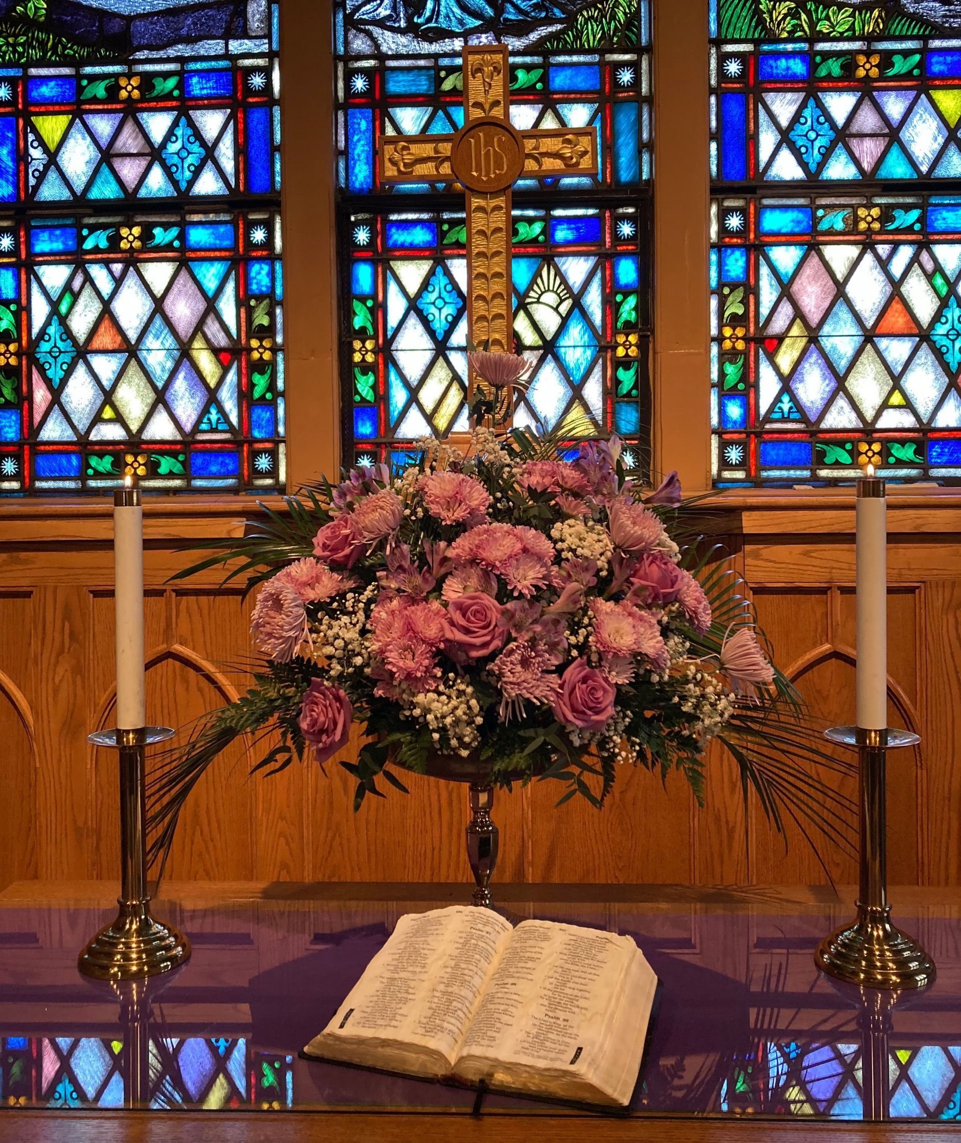 Altar with a cross, floral arrangement, candles, and open Bible in front of a stained-glass window.