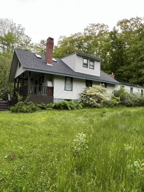 A white house with a porch and a chimney in the middle of a grassy field.