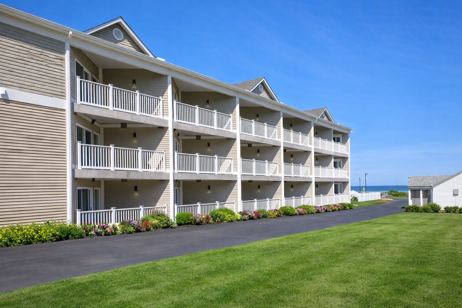 A multi-story beige hotel with tiered balconies faces a green lawn, a driveway, and the ocean under a clear blue sky.