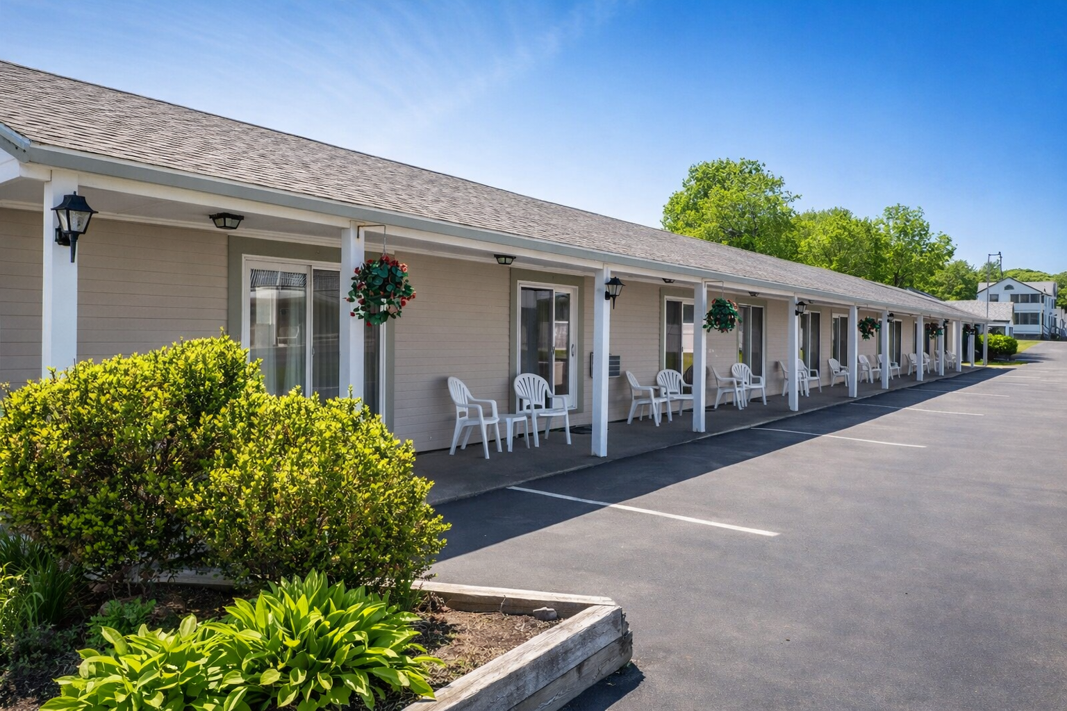 A row of hotel rooms with white porch columns, outdoor chairs, and a paved parking lot under a bright blue sky.