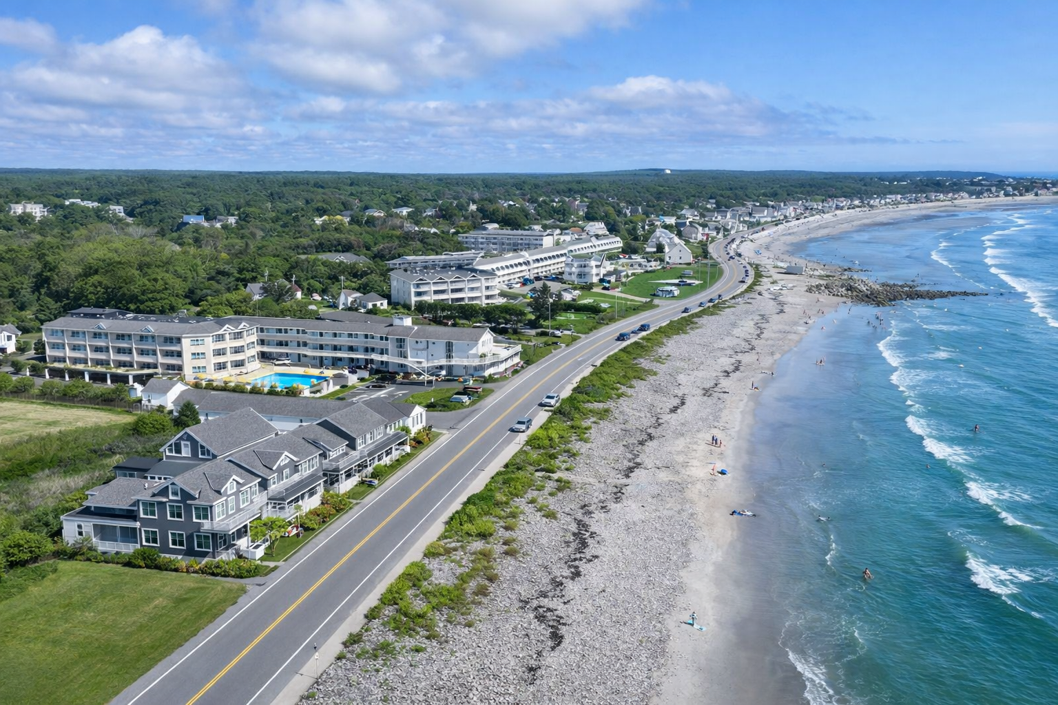 An aerial view of a coastal road separating a beach and ocean on the right from a hotel and lush landscape on the left.