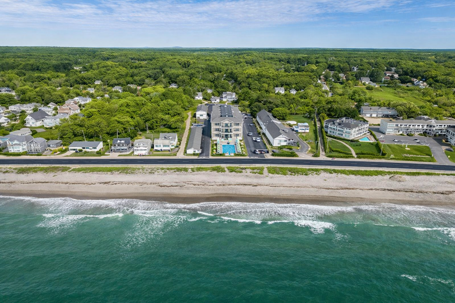 An aerial view of a multi-story beachside resort hotel with a pool, set against a shoreline and dense green forest.