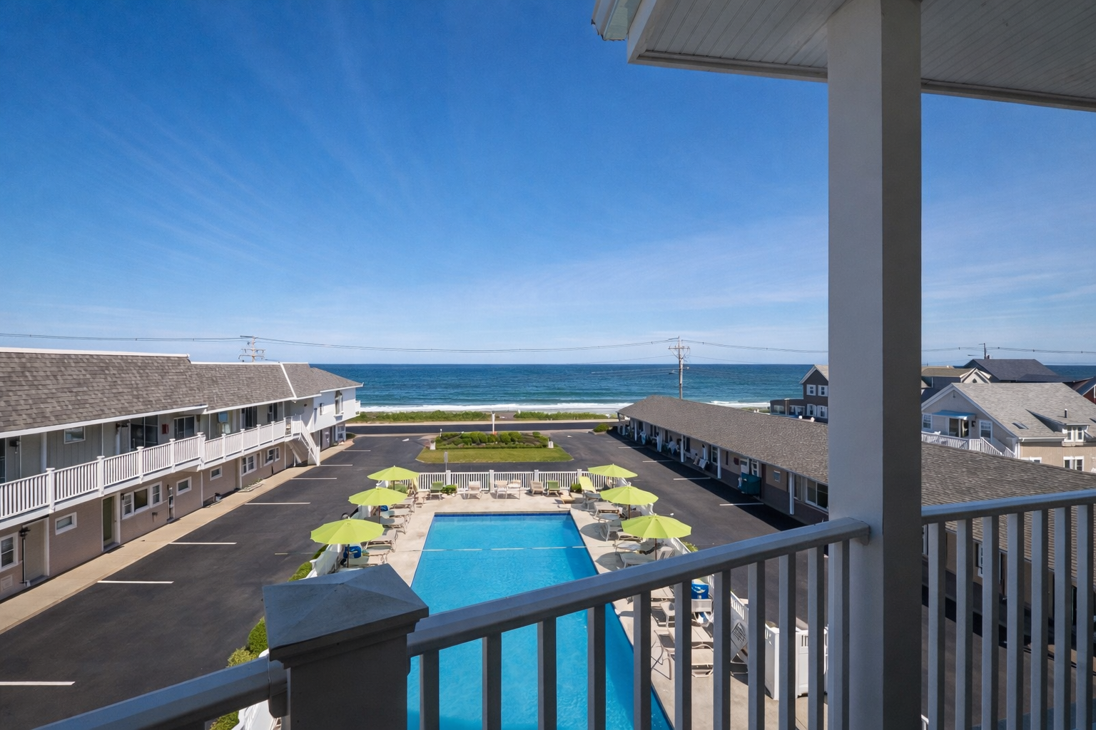 A high-angle view of an outdoor swimming pool surrounded by green umbrellas, flanked by beach resort buildings near the sea.
