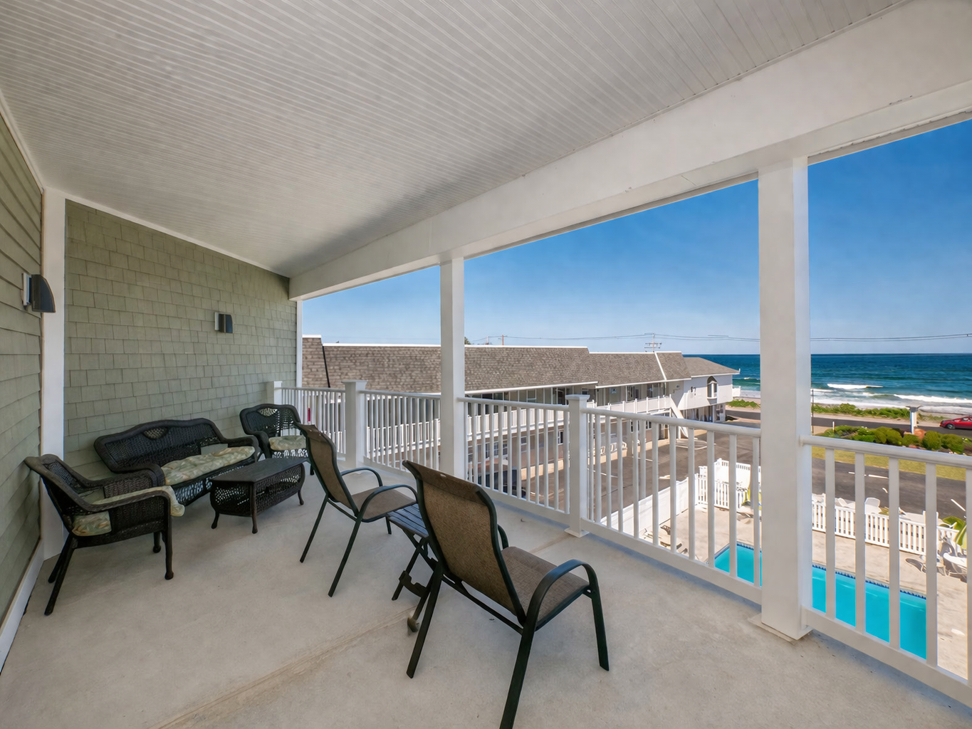 A covered balcony with outdoor seating overlooks a beach, ocean, and swimming pool on a sunny day.