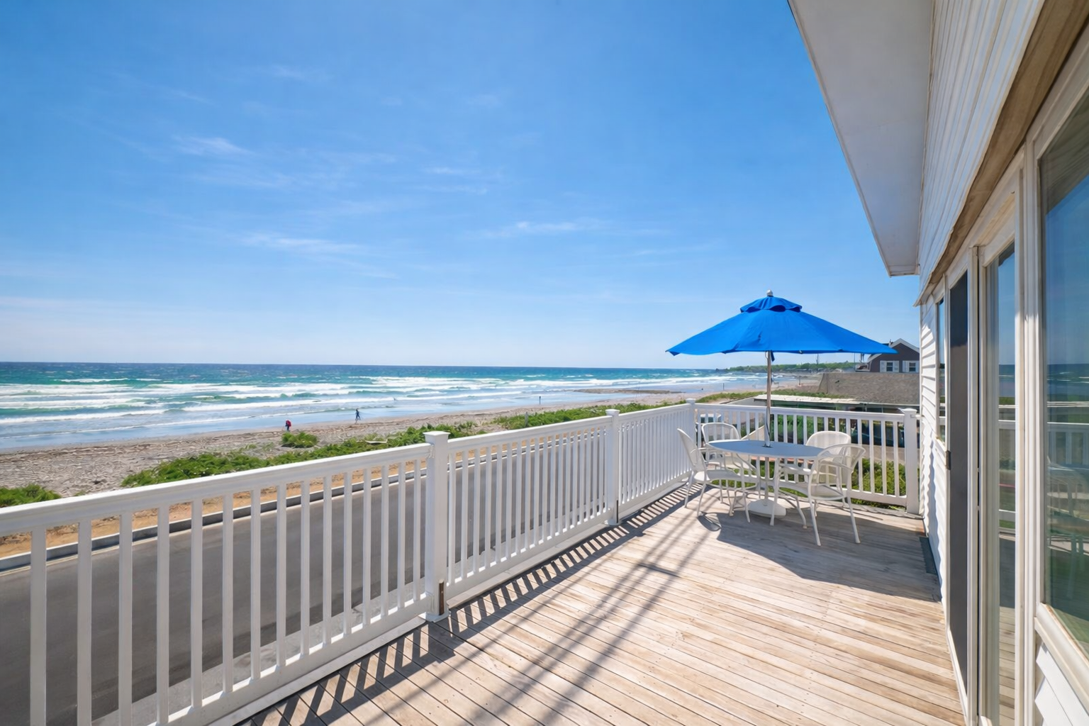 A wooden deck with a white railing overlooking a sandy beach and ocean, featuring a table with a bright blue umbrella.