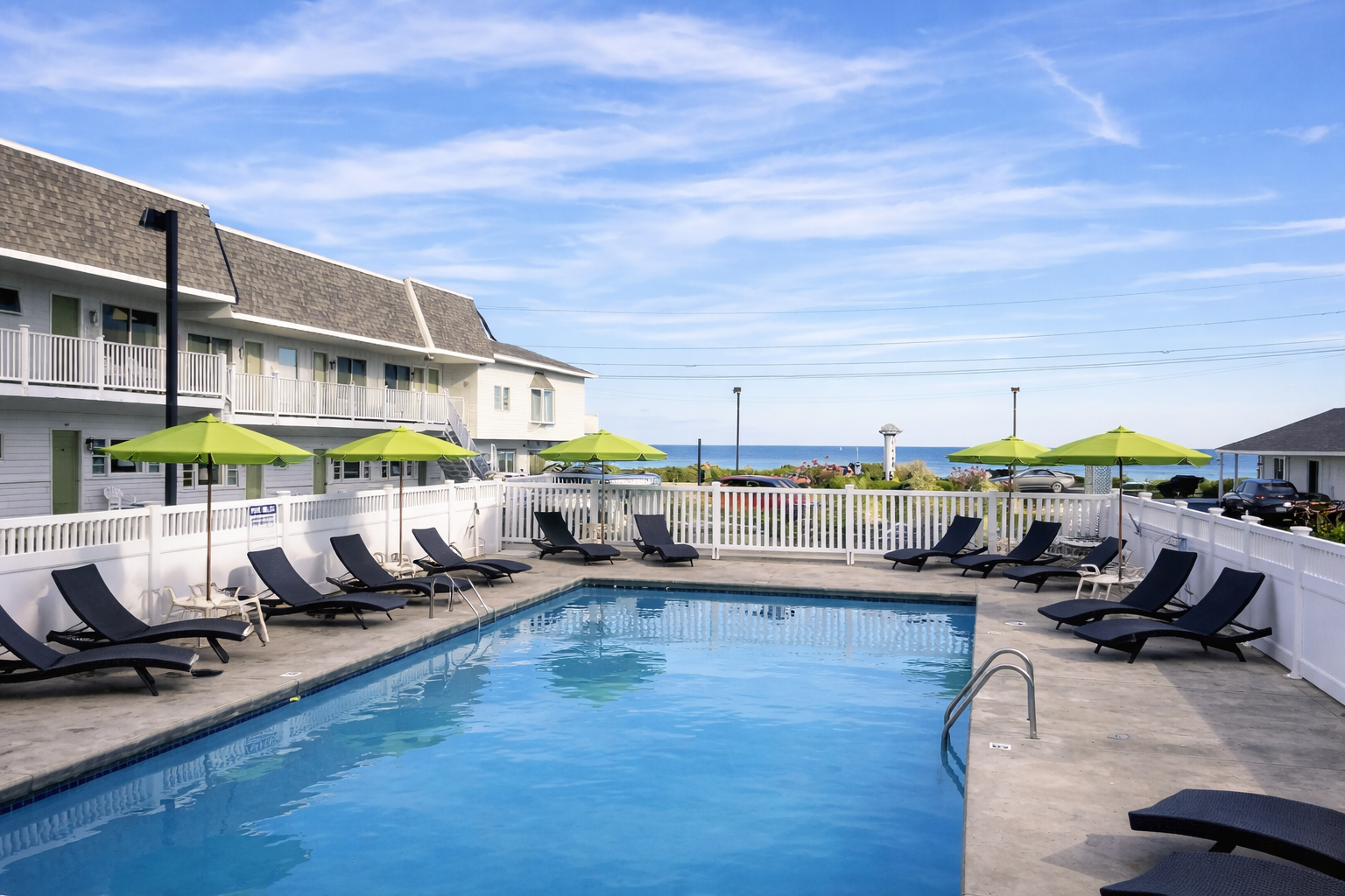 A swimming pool at a coastal hotel with lounge chairs, green umbrellas, and a white fence overlooking the ocean.