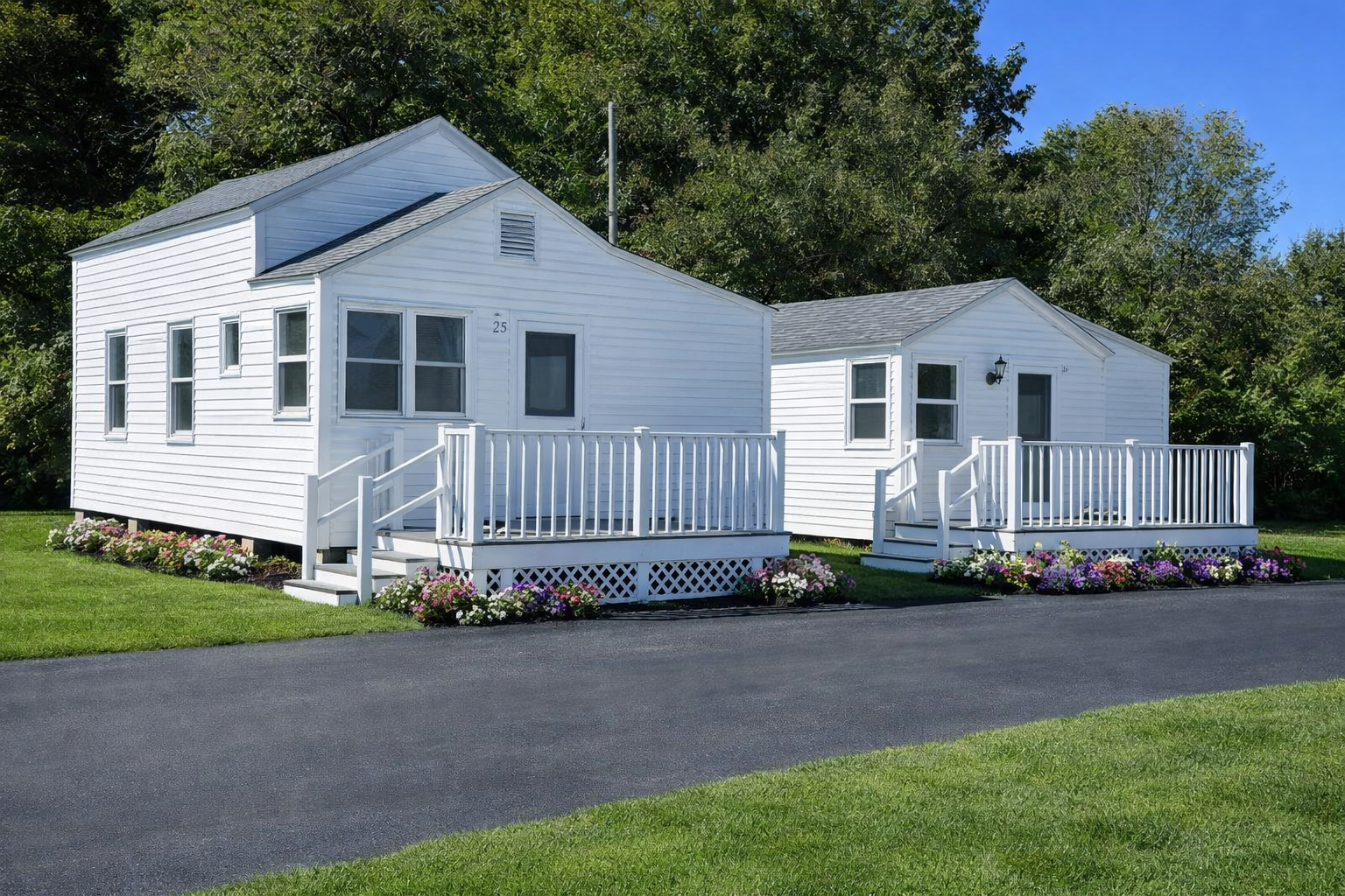 Two small white cottages with decks sit on a green lawn beside a paved driveway on a sunny day.
