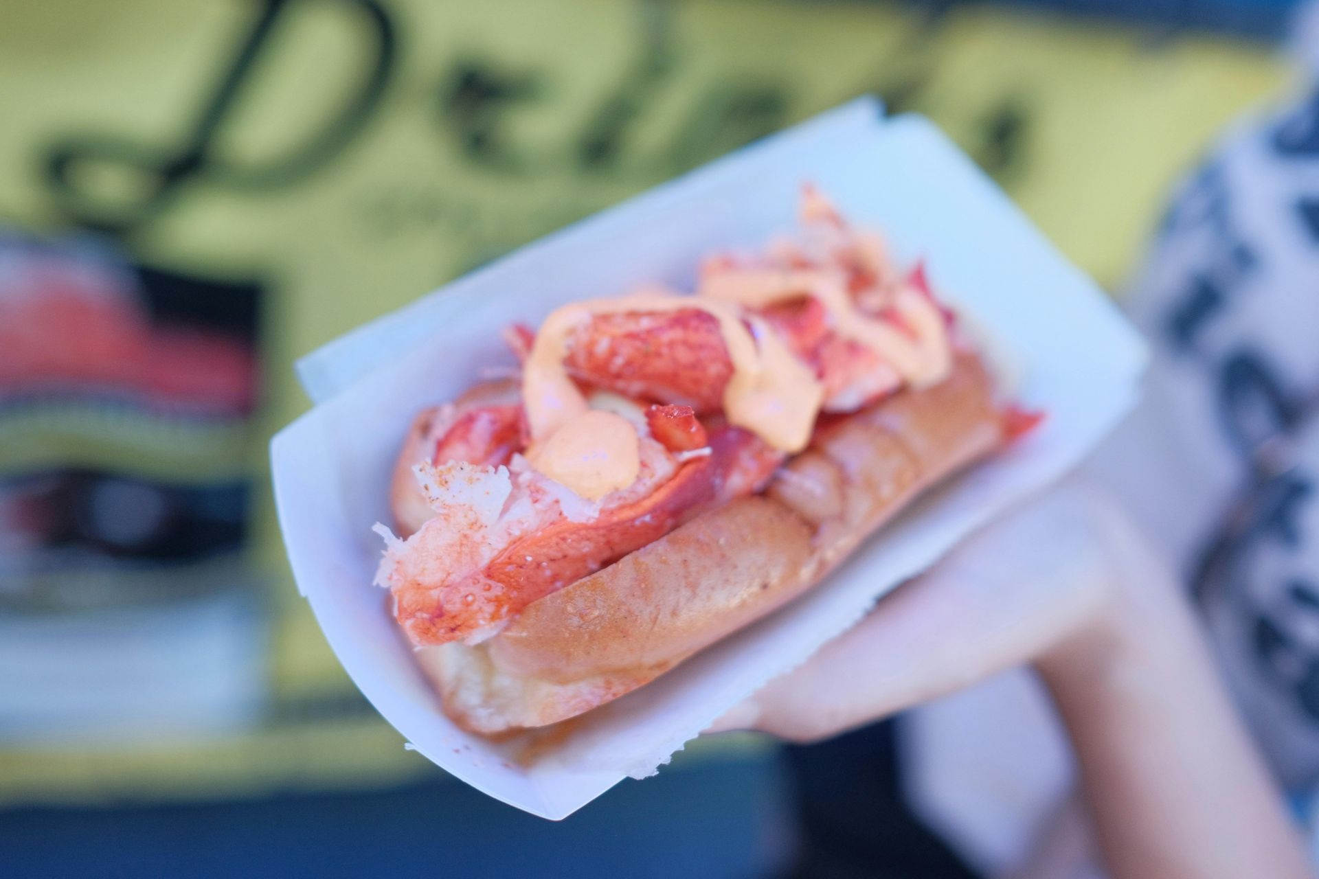 A hand holds a lobster roll topped with a drizzle of orange sauce in a paper tray against a blurry yellow background.
