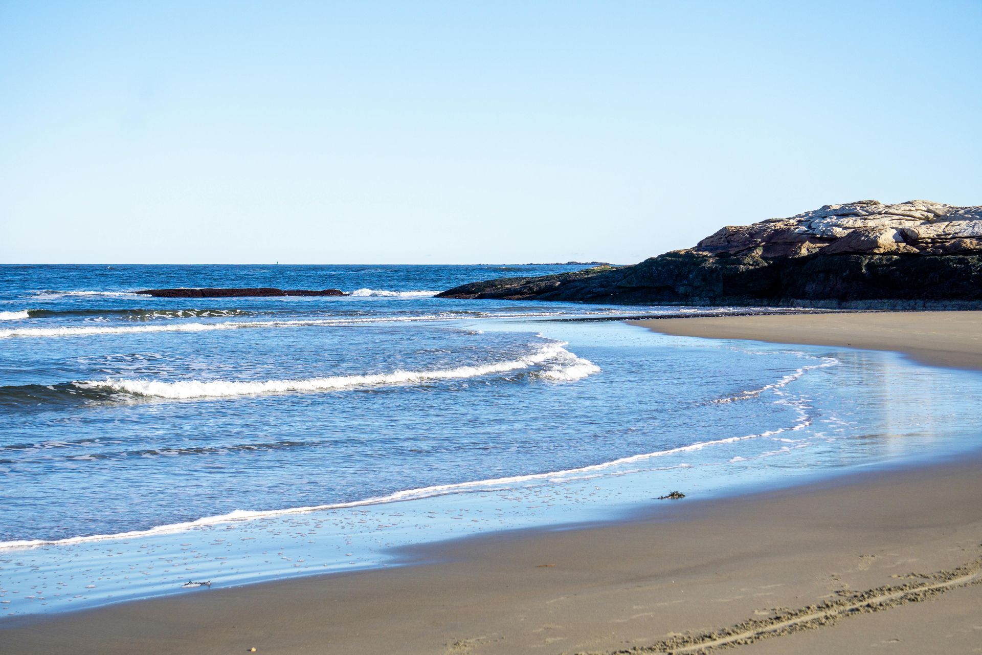 A sandy beach meets the ocean with gentle waves breaking on the shore, beside a rocky cliff under a clear blue sky.