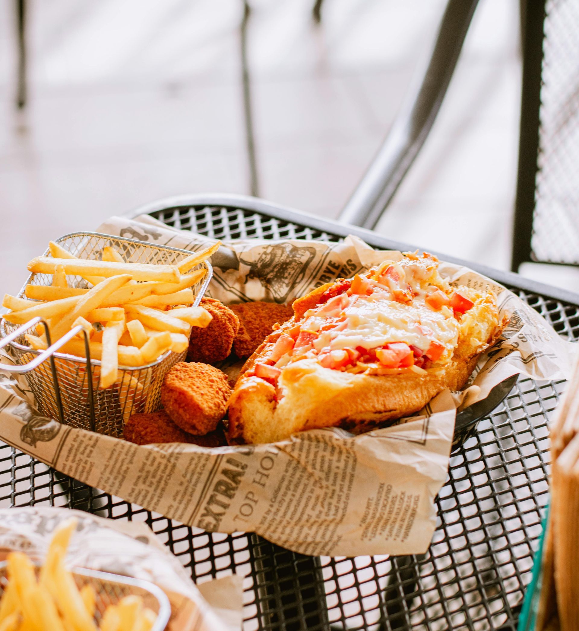 A basket lined with newspaper print holding french fries, round fritters, and a sandwich topped with diced tomatoes.