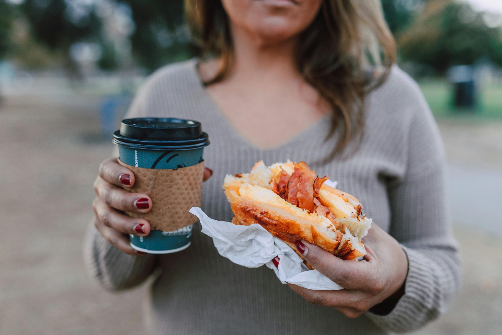 A person in a beige sweater holds a teal coffee cup and a bacon-filled sandwich in a paper wrapper outdoors.